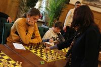 Stojąca kobieta wykonuje ruch białym gońcem na szachownicy, przed którą siedzi młody mężczyzna.