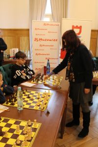 Stojąca kobieta trzyma białą figurę na szachownicy, przed którą siedzi chłopiec.