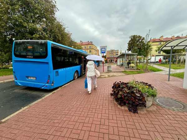 Obok niebieskiego autobusu kobieta ubrana na biało, z białą parasolką idąca w kierunku przystanku autobusowego.