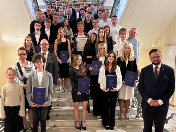 Grupa kilkudziesięciu dziewcząt, chłopców, kobiet i mężczyzn stoi na schodach patrząc przed siebie.