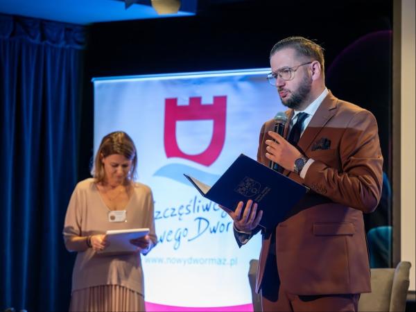 Mężczyzna w garniturze trzymający w prawej ręce otwartą teczkę mówi do mikrofonu trzymanego lewą ręką.