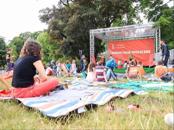 Grupa osób siedząca na leżakach i kocach przed sceną z banerem konsultacje społeczne.