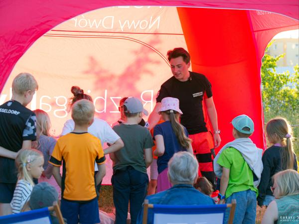 Grupa dzieci stojąca prze mężczyzną w czarnej koszulce i pomarańczowych spodniach.