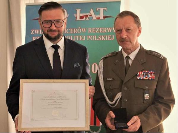 Obok mężczyzny w garniturze trzymającego obraz w ramce stoi mężczyzna w mundurze wojskowym.