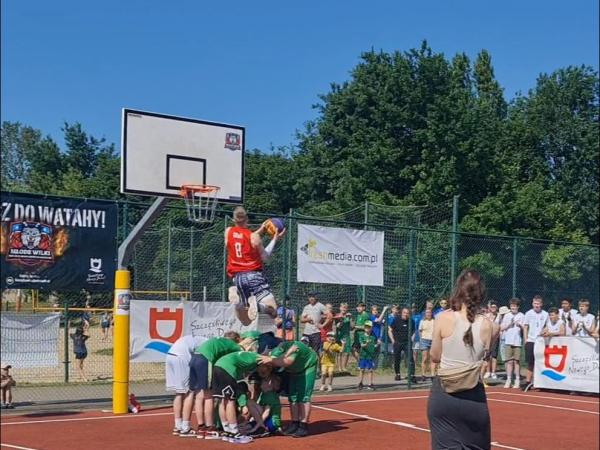 Mężczyzna z piłką do koszykówki w wyskoku nad grupą sześciu chłopców pochylonych do siebie.