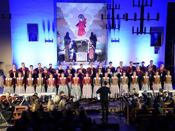 Śpiewające w kościele kobiety i mężczyźni ubrani w różnokolorowe stroje ludowe.