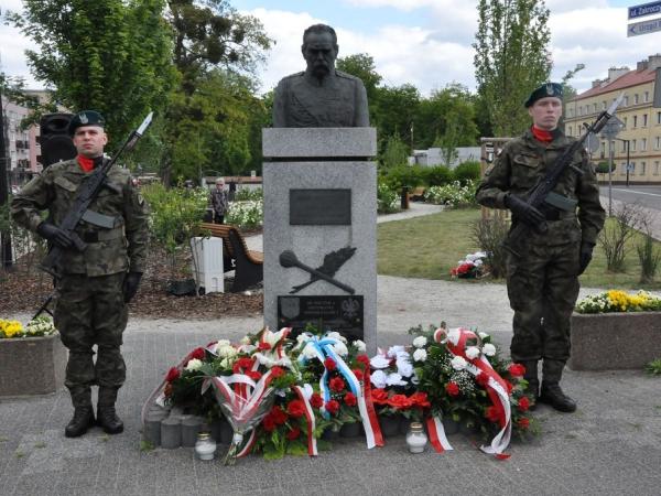 Dwóch żołnierzy z karabinami na piersiach stojących po obydwu stronach pomnika, poniżej wiązanki kwiatów.