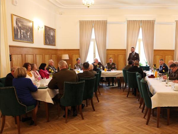 Kobiety i mężczyźni w mundurach siedzący za stołami, jeden mężczyzna w garniturze stoi za stołem.